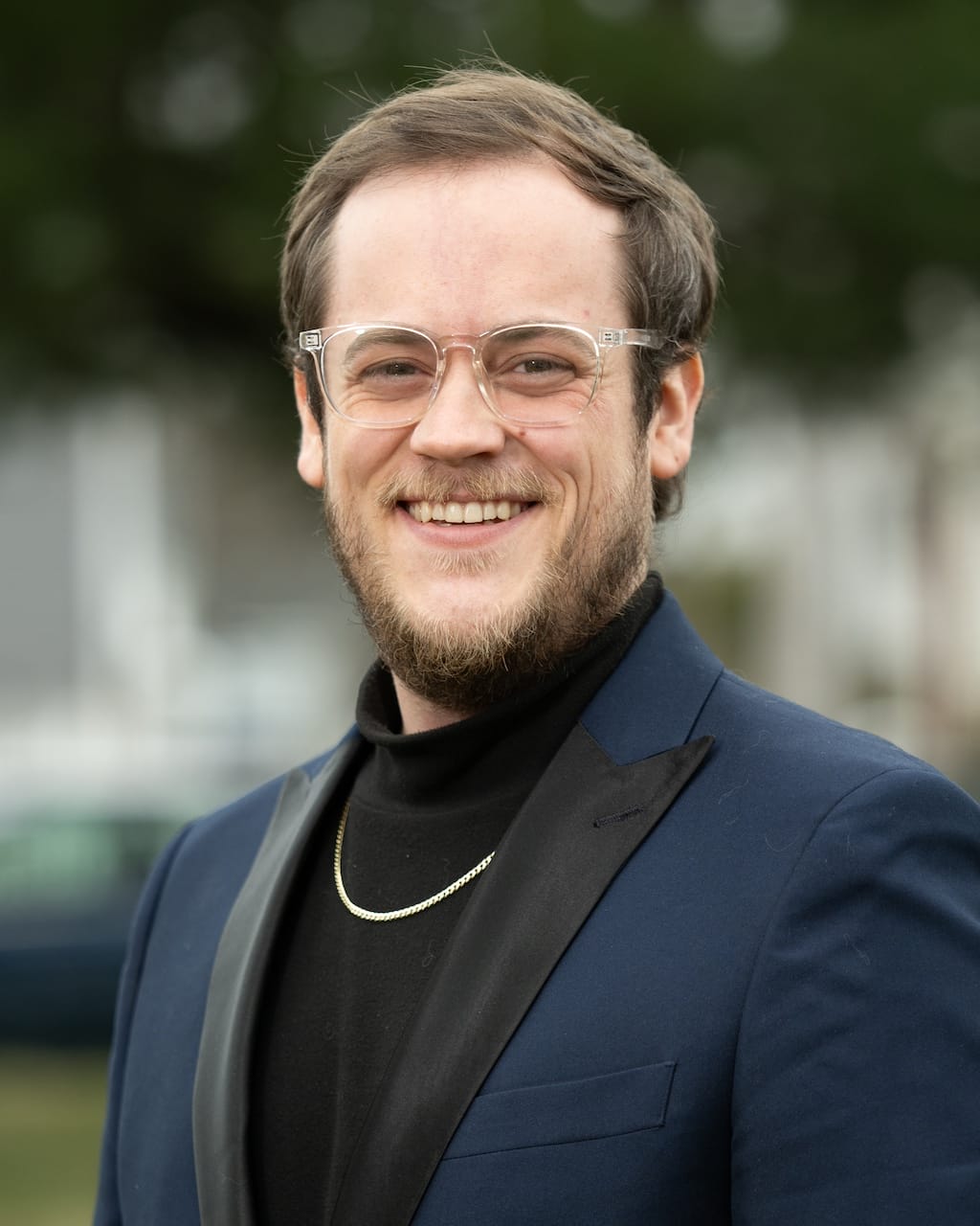 A man with glasses, a beard, and short hair, wearing a blue suit jacket over a black turtleneck and a gold chain, smiles at the camera. The background is blurred greenery.