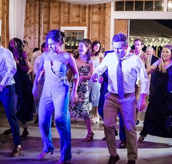 People dancing in a well-lit indoor venue with wooden walls and draped ceiling, wearing formal and semi-formal attire.