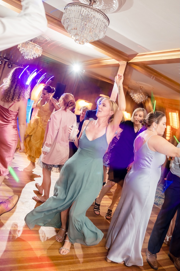 The Colony 1 A group of people in formal attire dance energetically under chandeliers at an indoor event, with colorful lighting effects in the background.