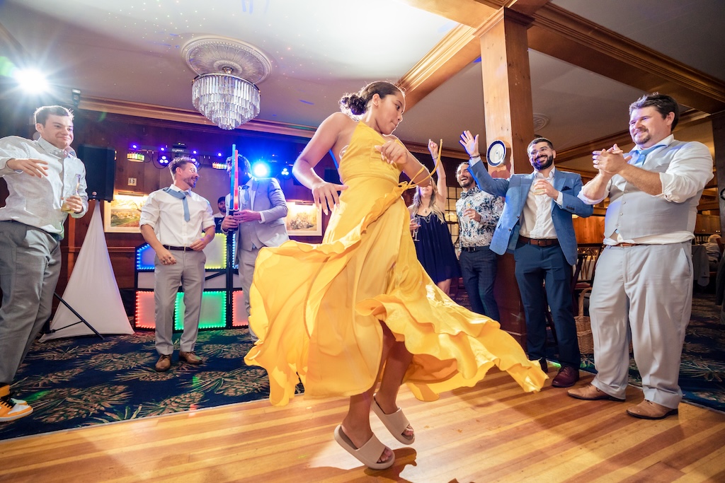 The Colony 2 A woman in a yellow dress dances energetically in the center of a wooden floor, surrounded by clapping and cheering people at a lively indoor event.