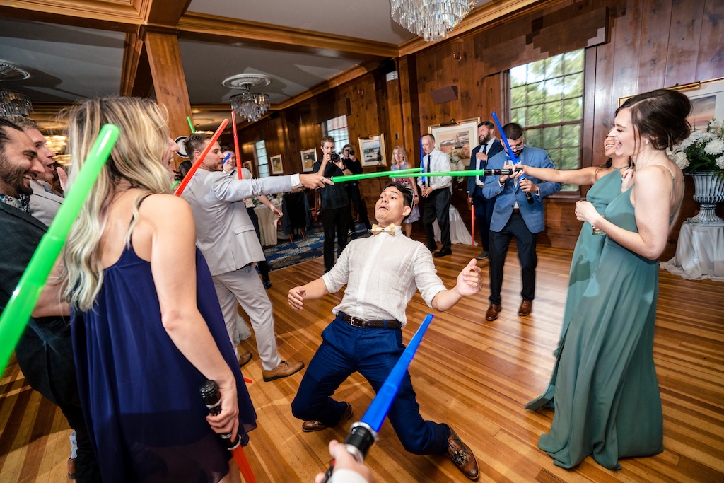 The Colony 3 A group of people in formal attire use toy lightsabers to form an arch while a man in a hat does the limbo beneath them at an indoor event.