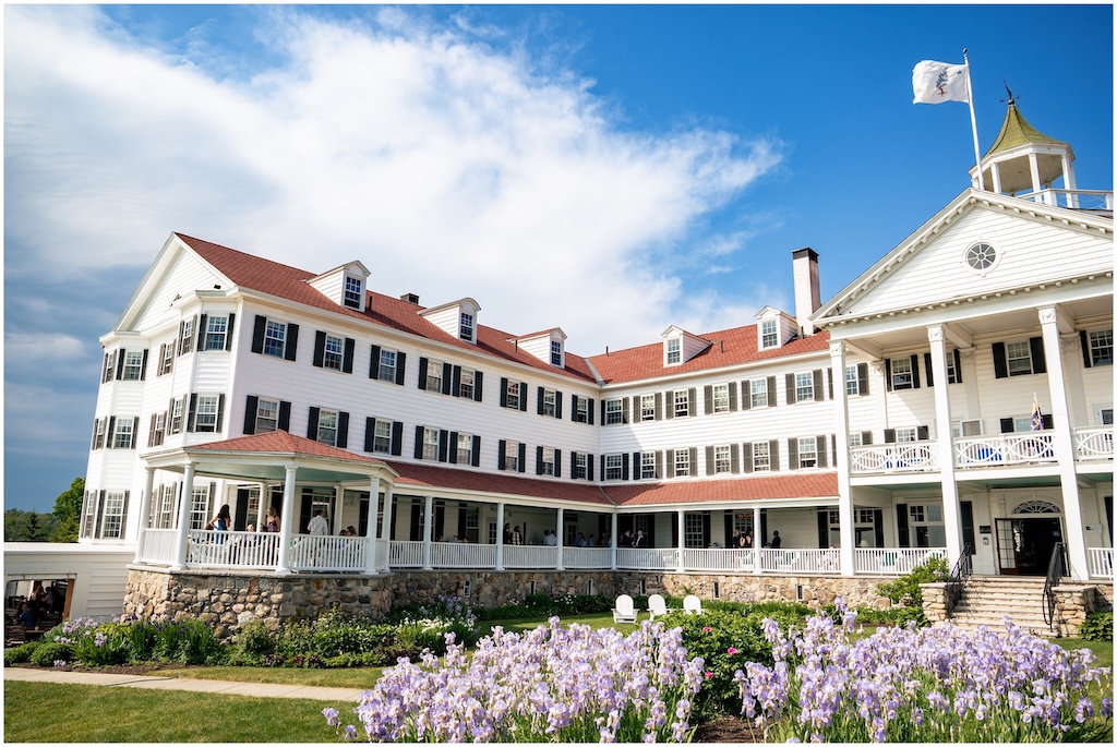 colony_hotel_wedding_photographers-_kennebunkport_wedding_photographer-_two_adventurous_souls-_060223_0037 A large white historic hotel with a red roof, wraparound porch, stone foundation, and flower garden in the foreground under a partly cloudy sky.