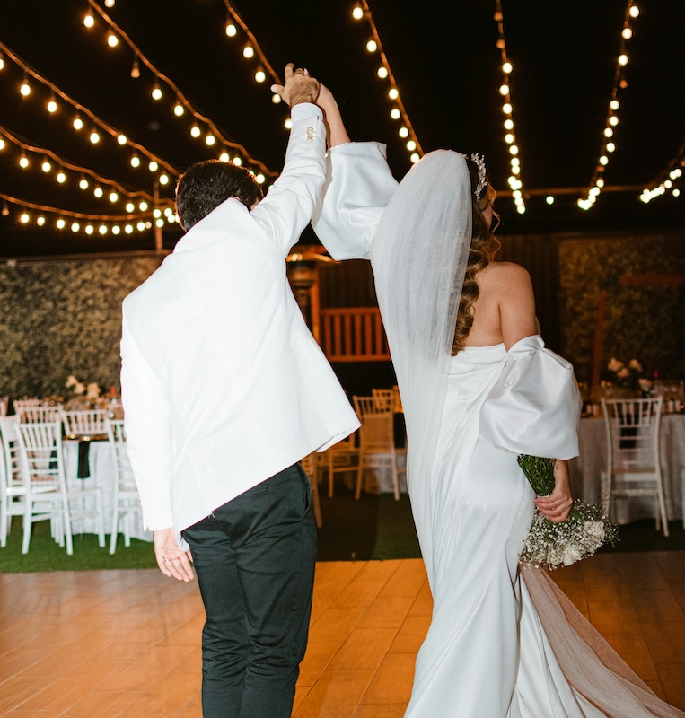 maine wedding reception djs A bride and groom, seen from behind, hold hands and raise their arms while walking under string lights at a wedding reception.