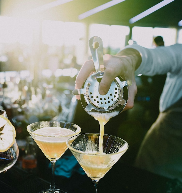the colony hotel coacktail hour A bartender pours a yellow cocktail through a strainer into two martini glasses at a bar with bright lighting.