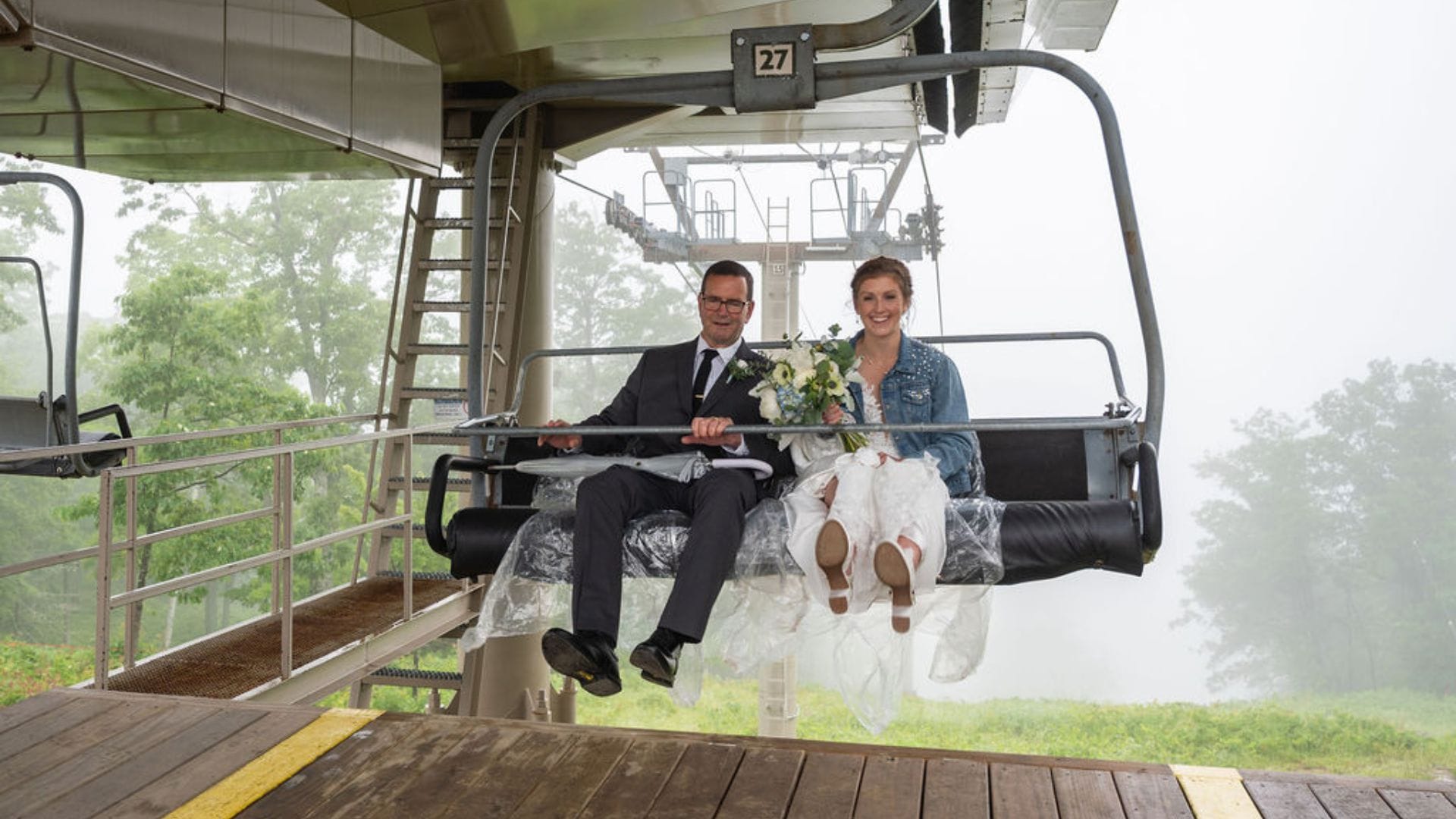 A bride and her dad sit on a ski lift at a mountaintop New Hampshire wedding, the bride holding a bouquet and wearing a denim jacket. Trees and fog are visible in the background.