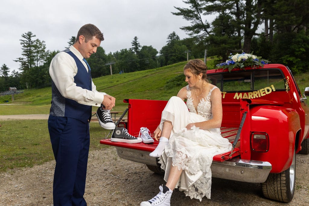 A bride and groom sit on the tailgate of a red pickup truck with a "Married!" sign, putting on matching black-and-white sneakers after their mountaintop New Hampshire wedding.