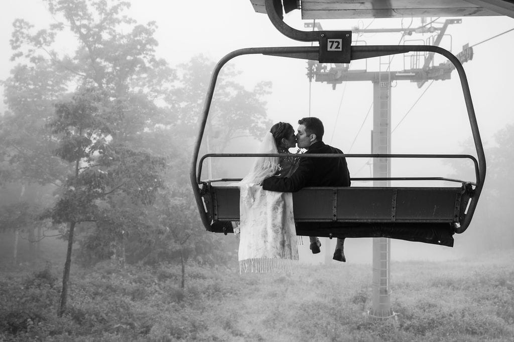 A bride and groom sit together on a ski lift, embracing as they ascend through a misty, tree-lined landscape—capturing the magic of a mountaintop New Hampshire wedding.