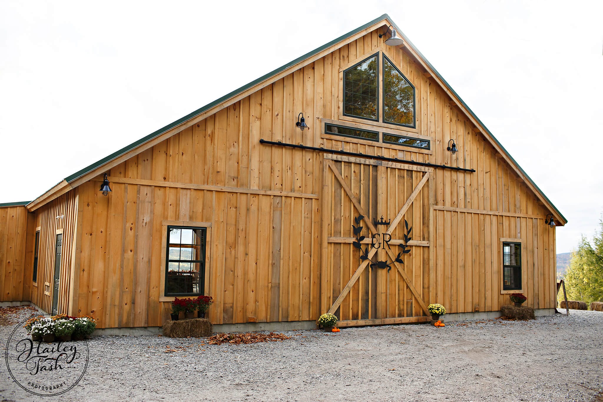 A large wooden barn with light brown siding, double sliding doors, several windows, and decorative hay bales and potted flowers placed outside.