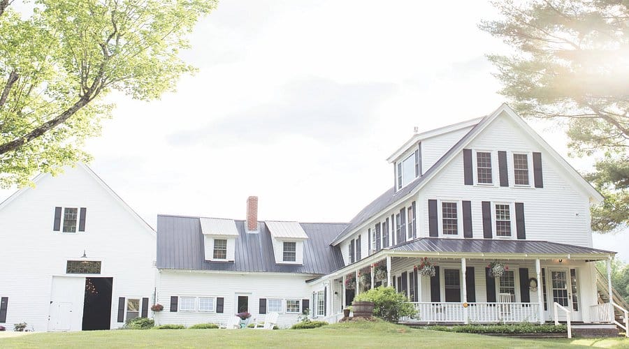 Large white farmhouse with black shutters and a wraparound porch, set on a well-kept lawn with trees and bright sunlight in the background.