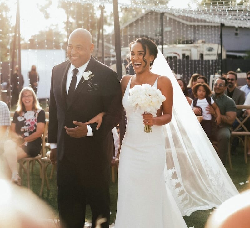 A bride in a white dress and holding a bouquet walks down the aisle with a man in a suit during an outdoor wedding ceremony.