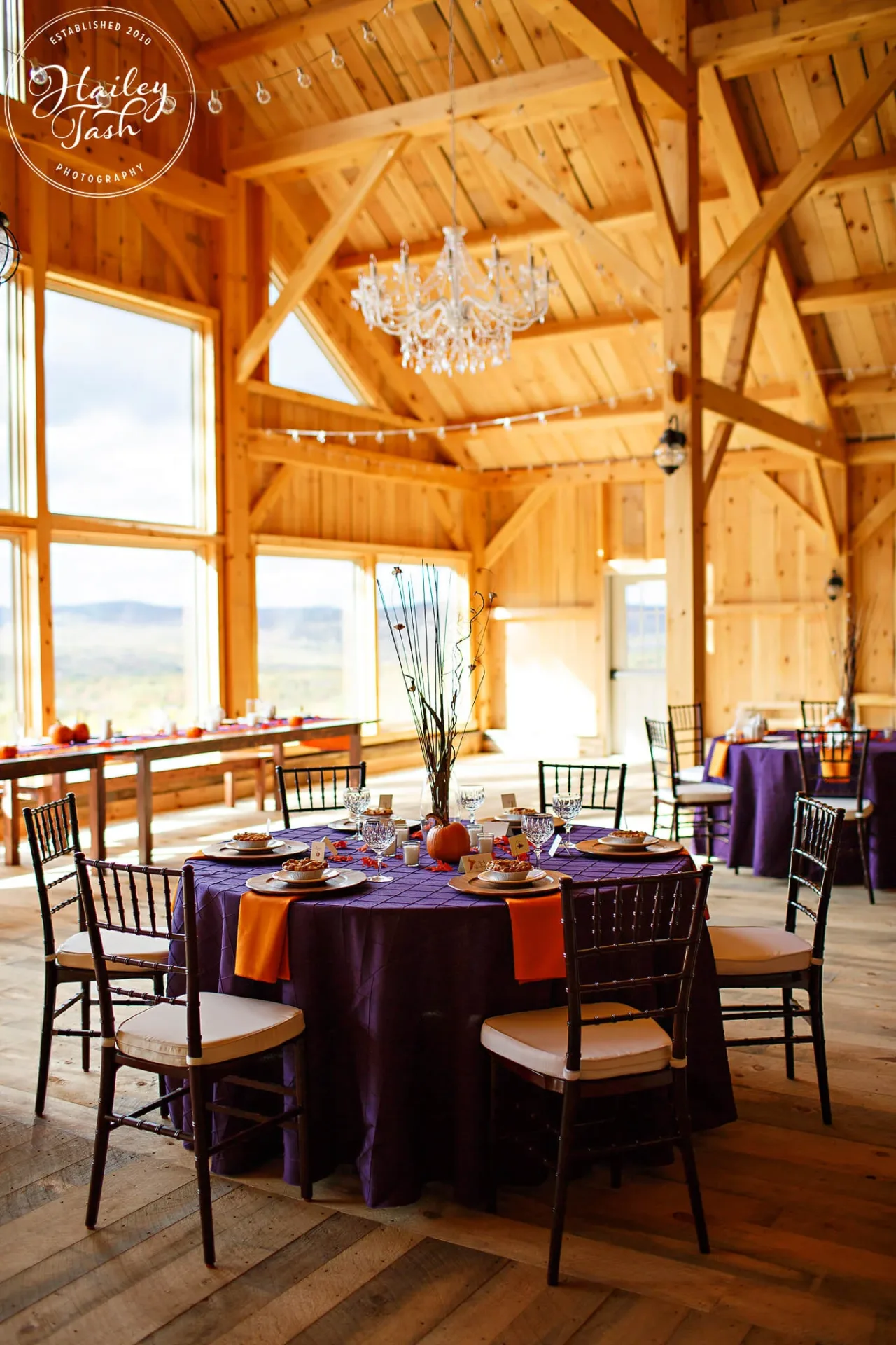A round table with purple tablecloth, orange napkins, and simple centerpiece is set in a wooden banquet hall with large windows and a chandelier.