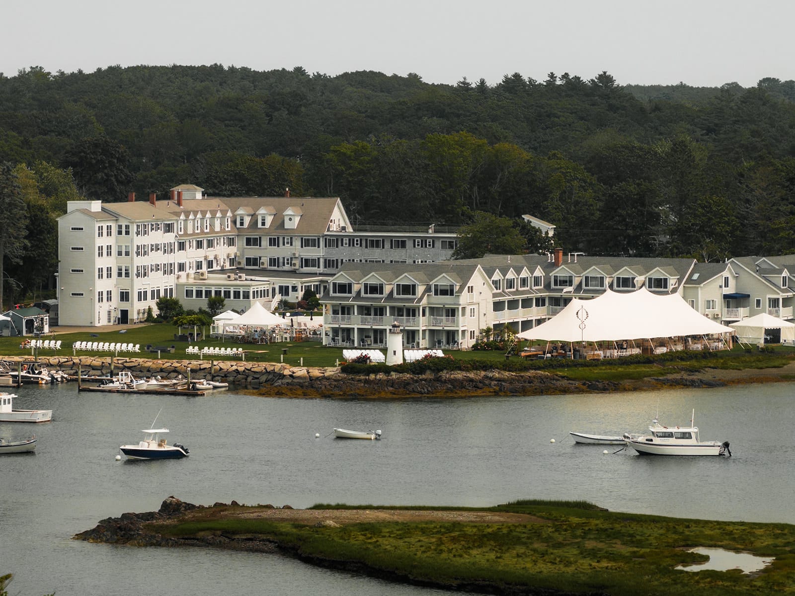 A large waterside resort with white buildings, a tented event area, and several boats docked along the shoreline, surrounded by trees.