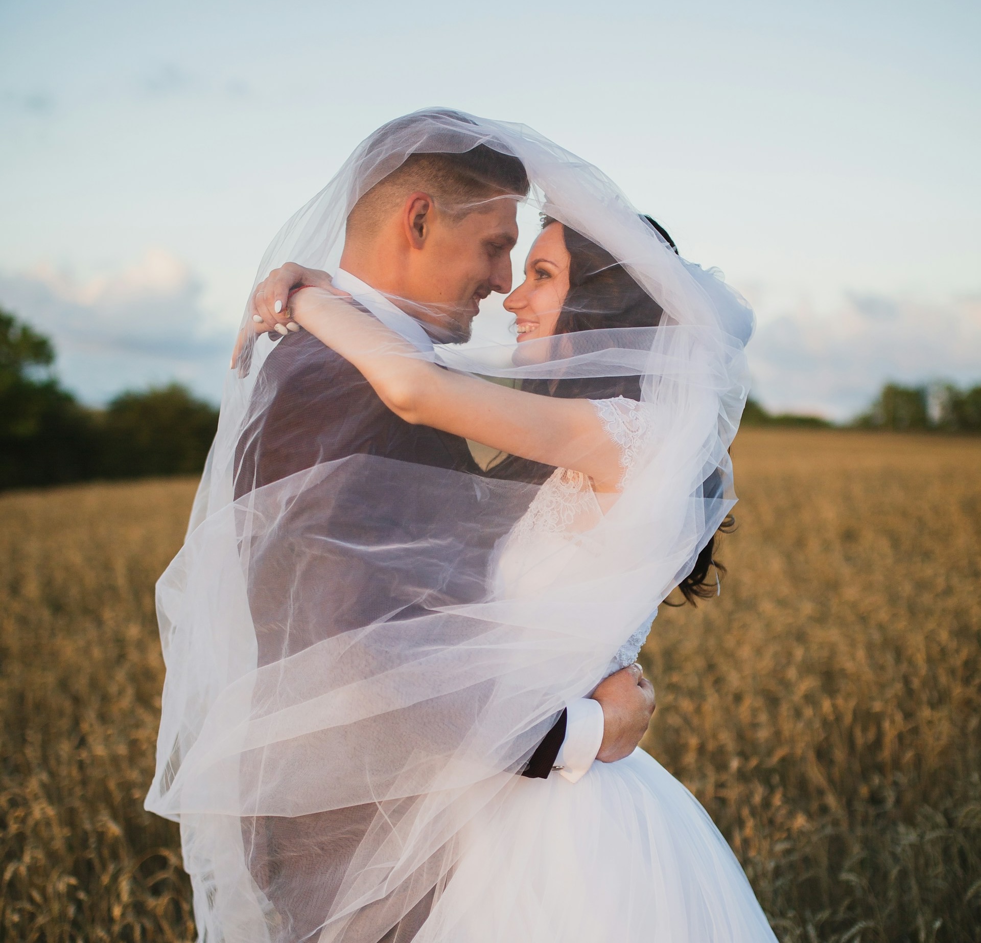 north conway, NH weddings A bride and groom embrace in a field, wrapped in a flowing wedding veil, looking into each other's eyes at sunset.