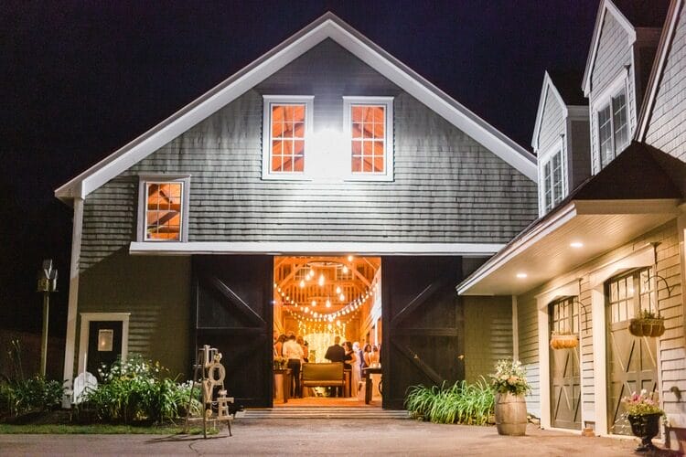 A barn at night with open doors reveals people inside under string lights, with a “LOVE” sign and plants decorating the entrance.