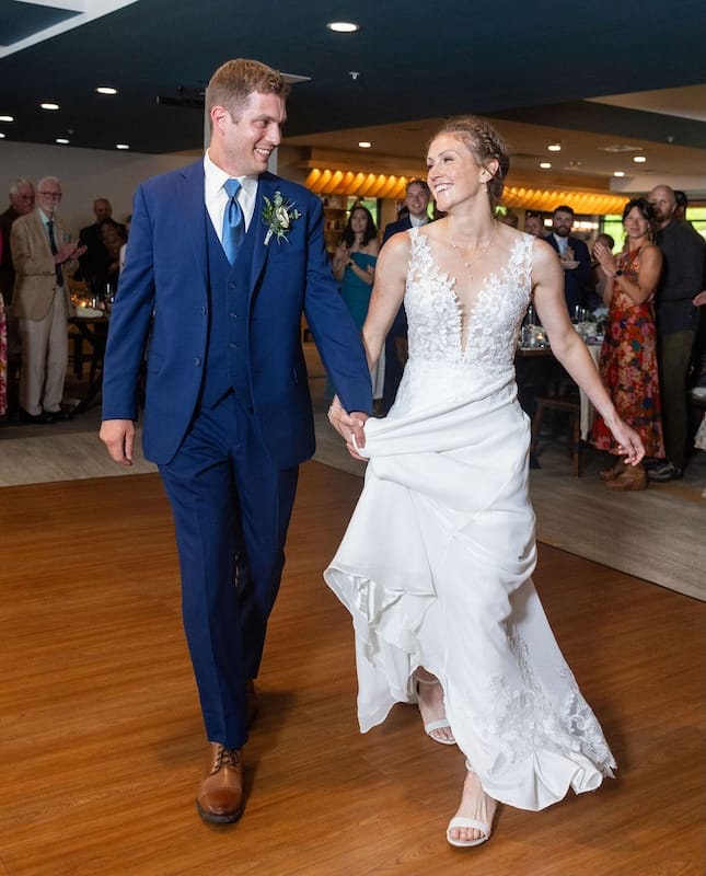 A bride and groom smile and hold hands while walking together indoors at their wedding reception, with guests watching in the background.