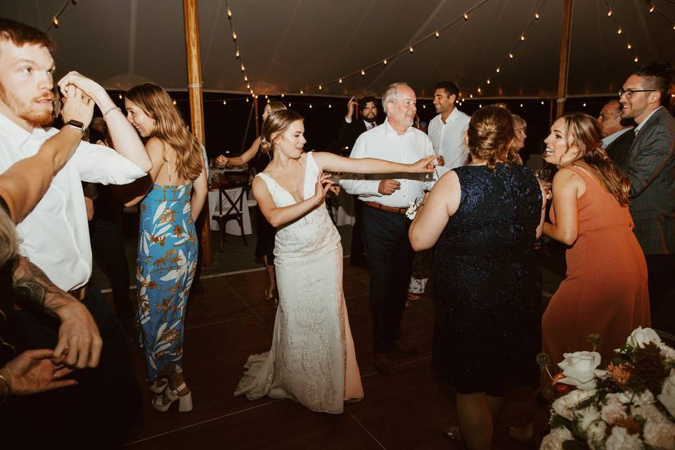 People dance and celebrate on a wooden floor under string lights at a wedding reception, with the bride in the center wearing a white dress.