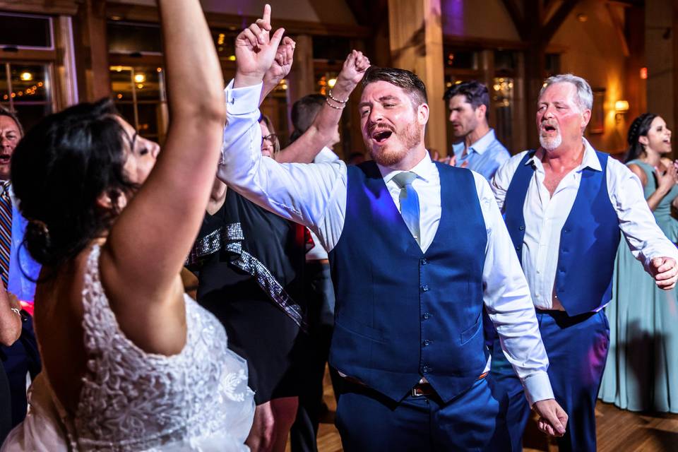 A group of people dressed in formal attire dance enthusiastically at what appears to be a wedding reception, perhaps celebrating after learning How Much Does a Wedding DJ Cost in Maine vs. New Hampshire.