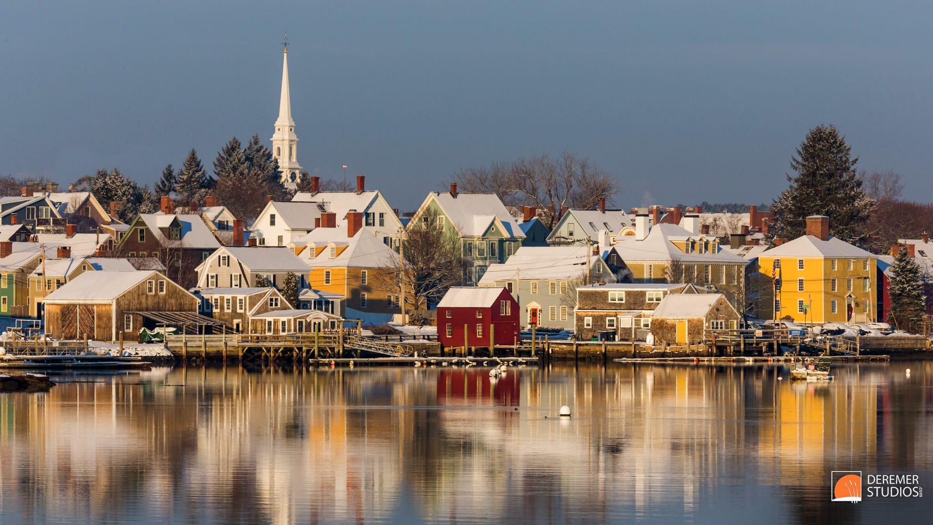 2013-12-fine-nh-winter-sunrise-20-portsmouth-nh-north-church-waterfront Snow-covered houses and buildings line a calm waterfront, with a white church steeple in the background under a clear sky.