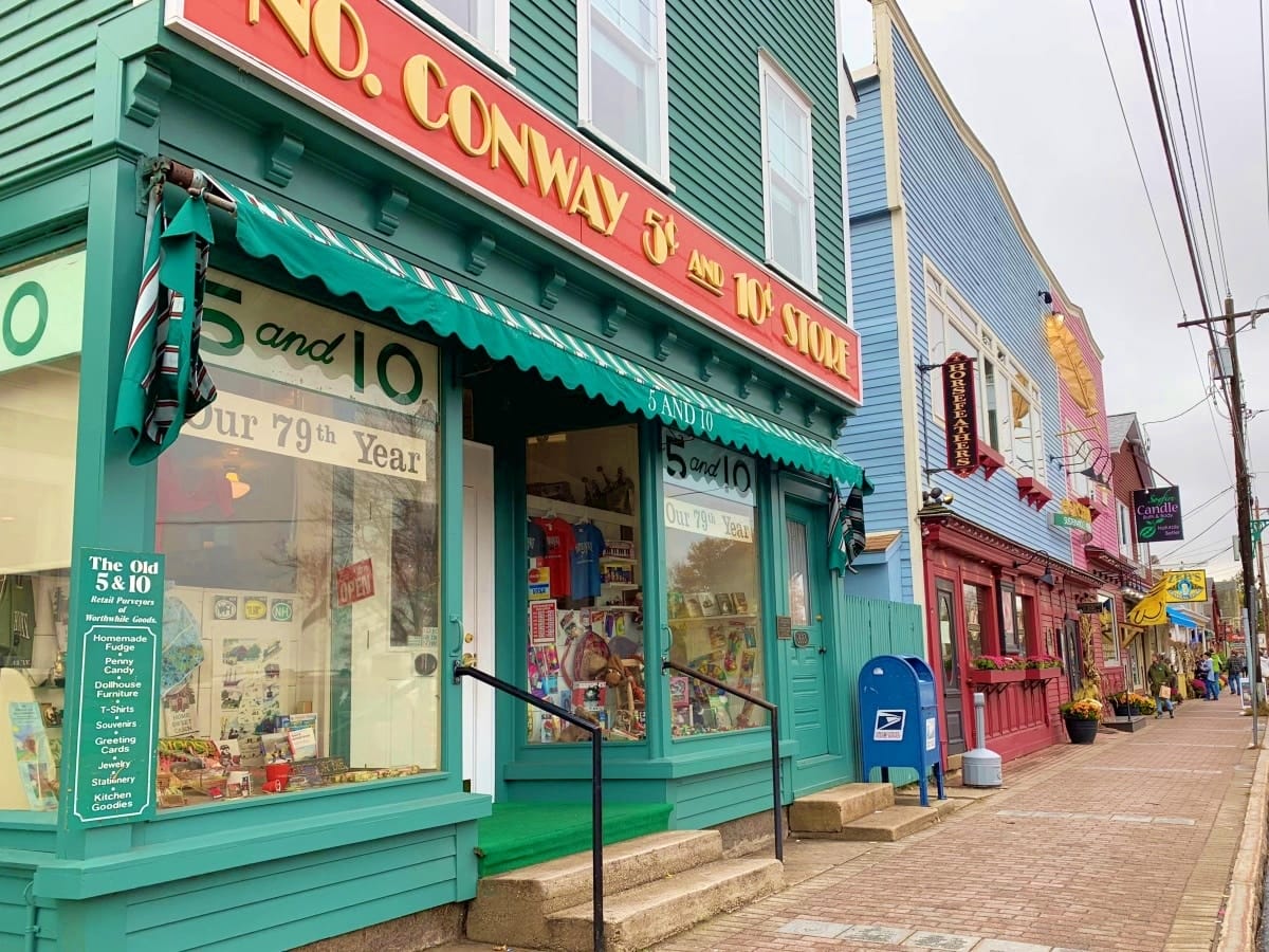 IMG_0477-1 Colorful storefronts line a small town street, with a green awning above a shop labeled “No. Conway 5¢ and 10¢ Store” and a blue mailbox on the sidewalk.
