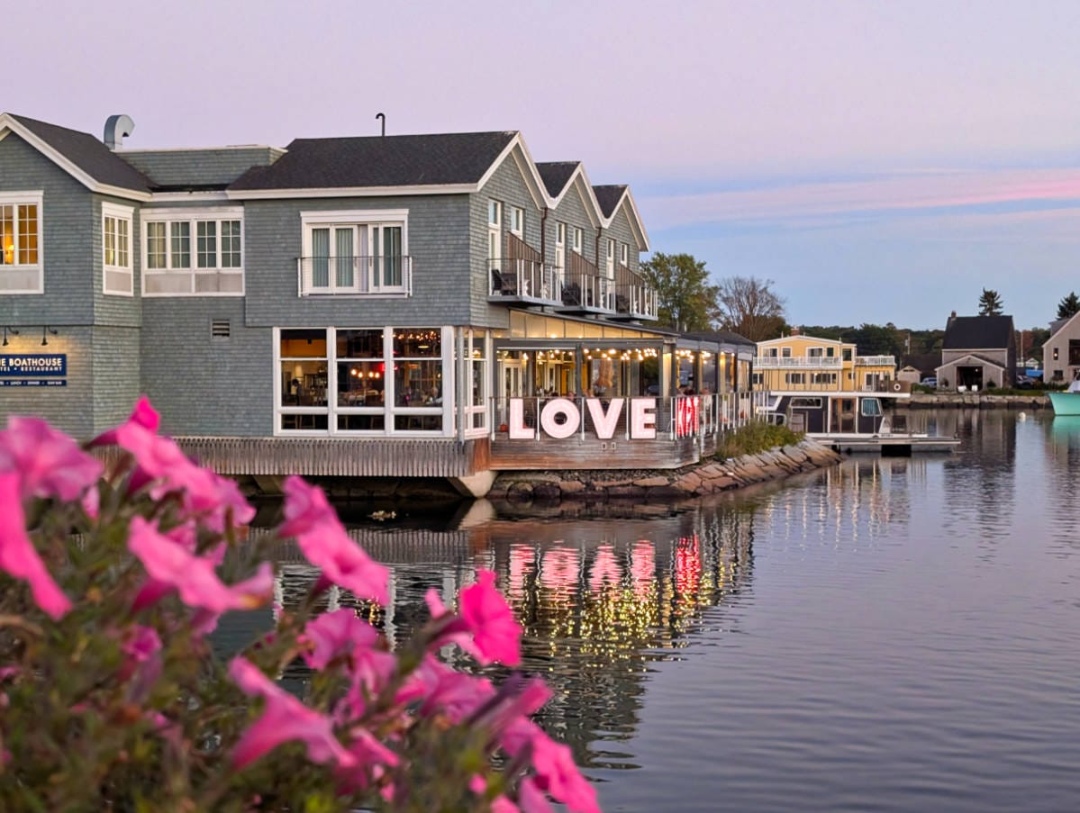 Waterfront building with large illuminated "LOVE" sign, pink flowers in the foreground, and calm water reflecting the scene at dusk.