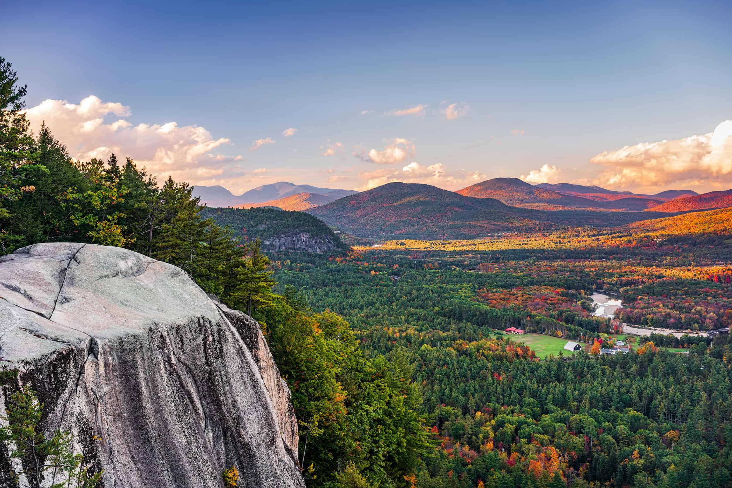 North Conway View from a rocky cliff overlooking a forested valley with distant mountains, scattered houses, a winding river, and a colorful sky at sunset.