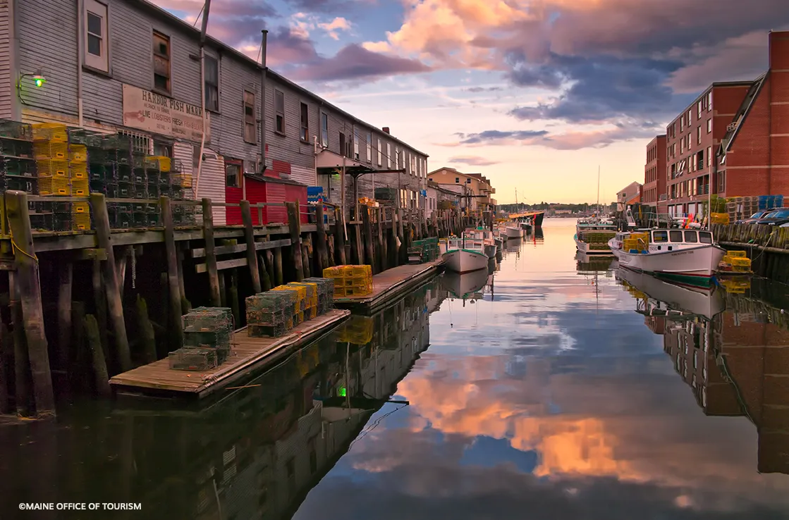 Lobster traps stacked along wooden docks, fishing boats moored in a narrow harbor, and colorful sunset clouds reflected in calm water.