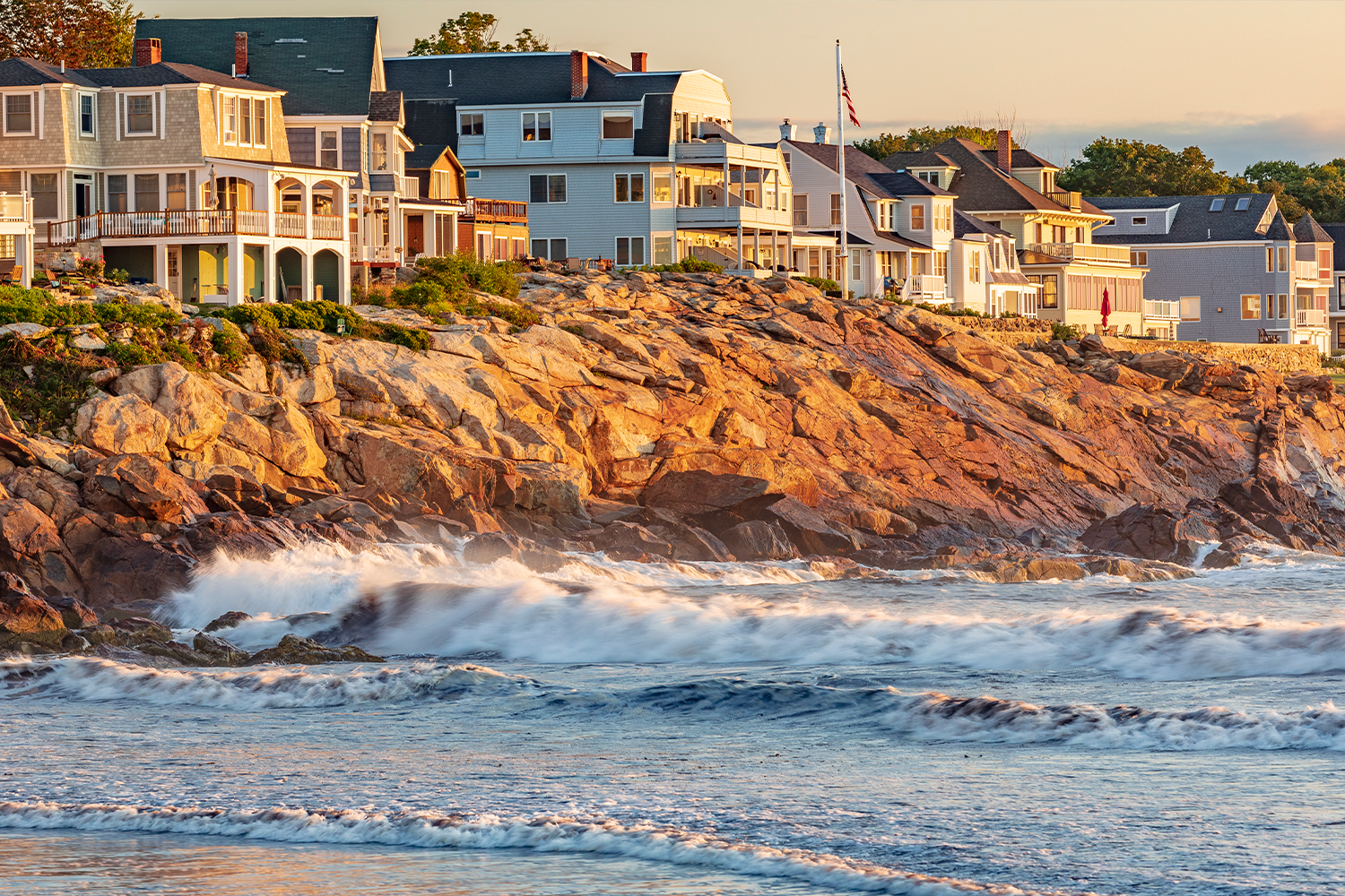 York-Maine.png Coastal houses sit atop rocky cliffs as ocean waves crash below, bathed in warm sunlight.