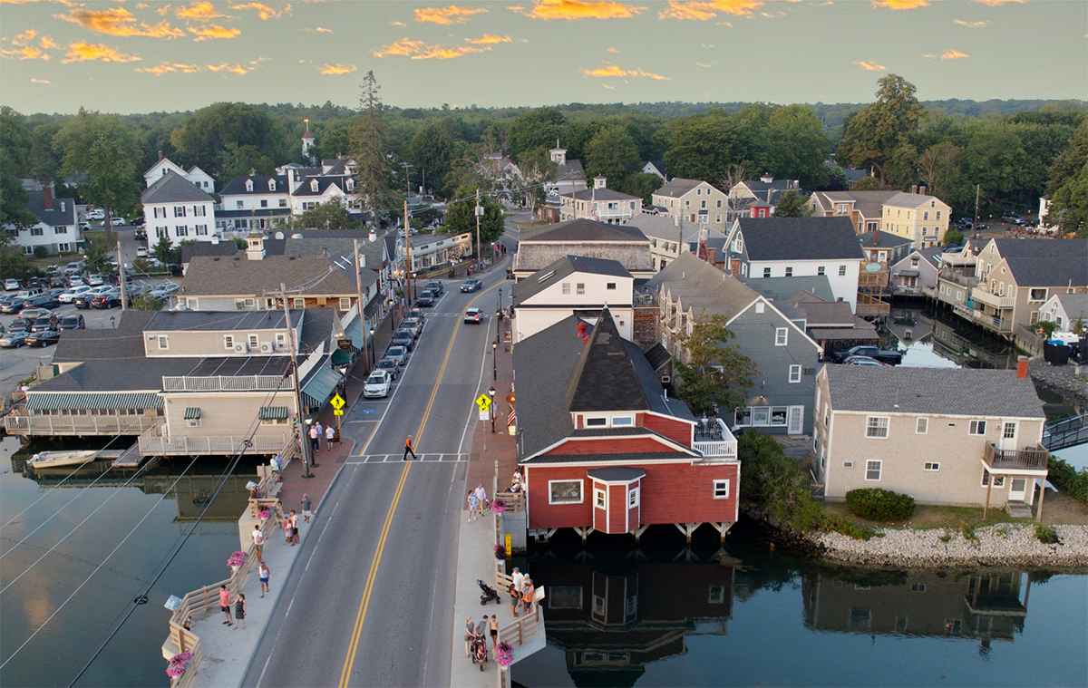 Aerial view of a small town with a road crossing over water, surrounded by buildings and trees under a partly cloudy sky at sunset.