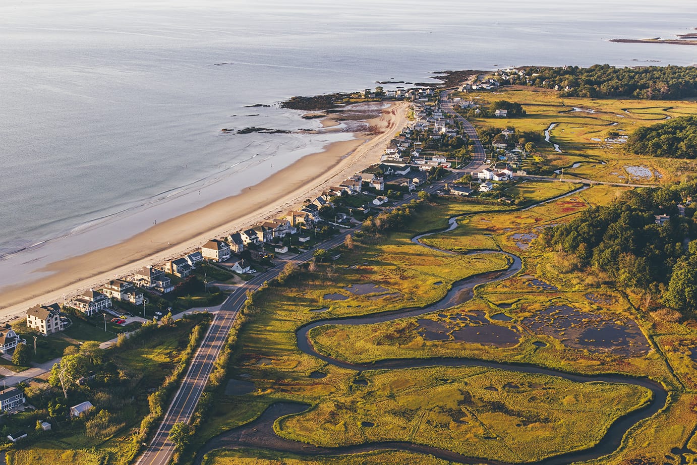 Aerial view of a coastal town with houses lined along a sandy beach, winding marshlands, and a road running parallel to the shoreline.