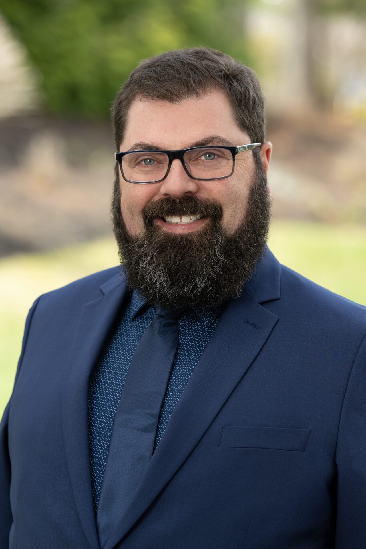 A man with dark hair, glasses, and a full beard is wearing a navy blue suit and tie, standing outdoors with a blurred background.