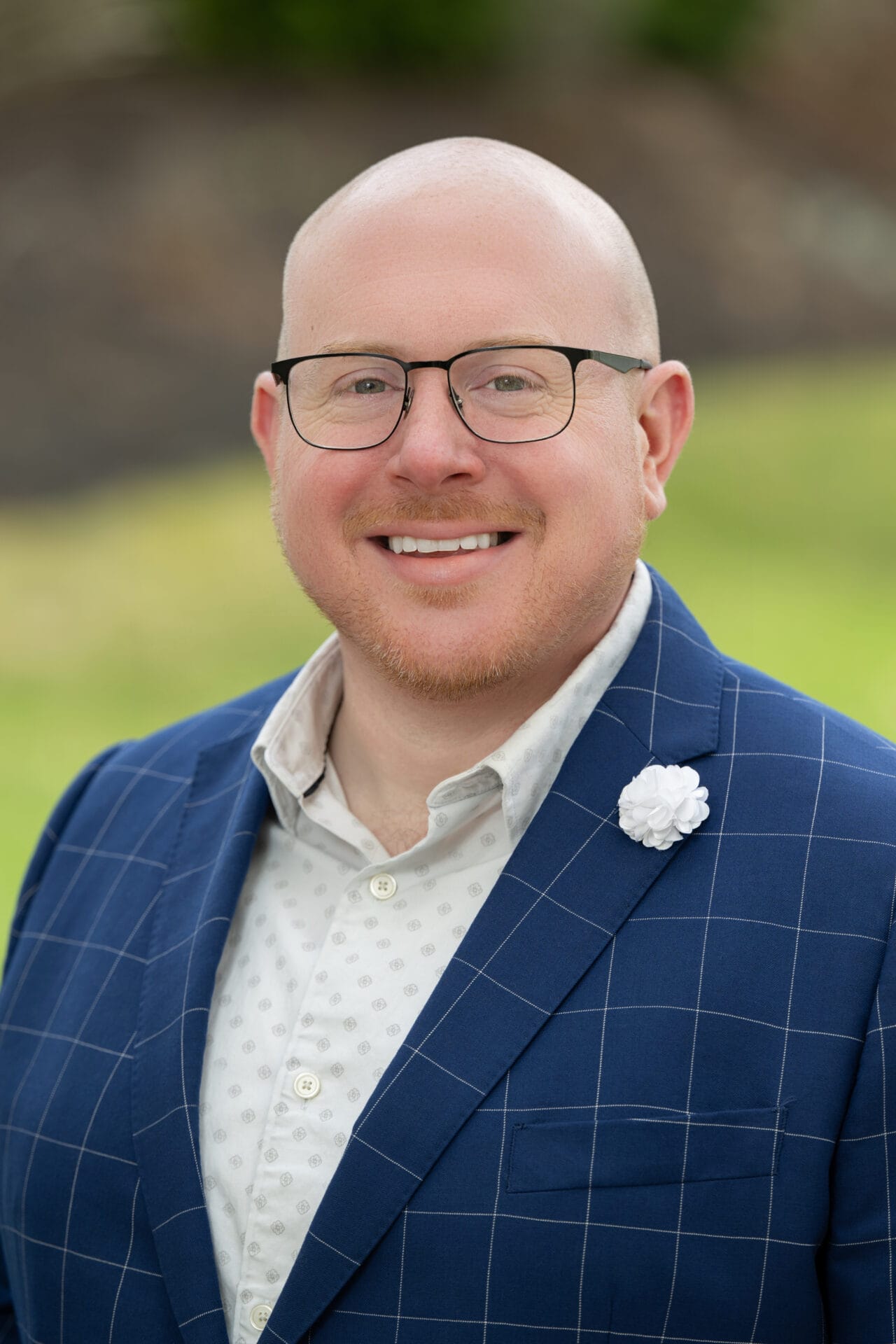 A smiling person with glasses and a bald head wears a blue checked suit, white shirt, and a white flower pin, standing outdoors with greenery in the background.