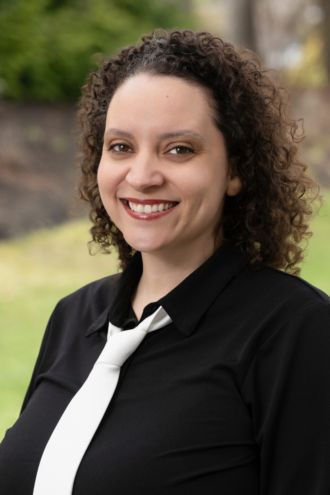 A person with curly hair, wearing a black shirt and white tie, smiles outdoors with greenery and a blurred background.