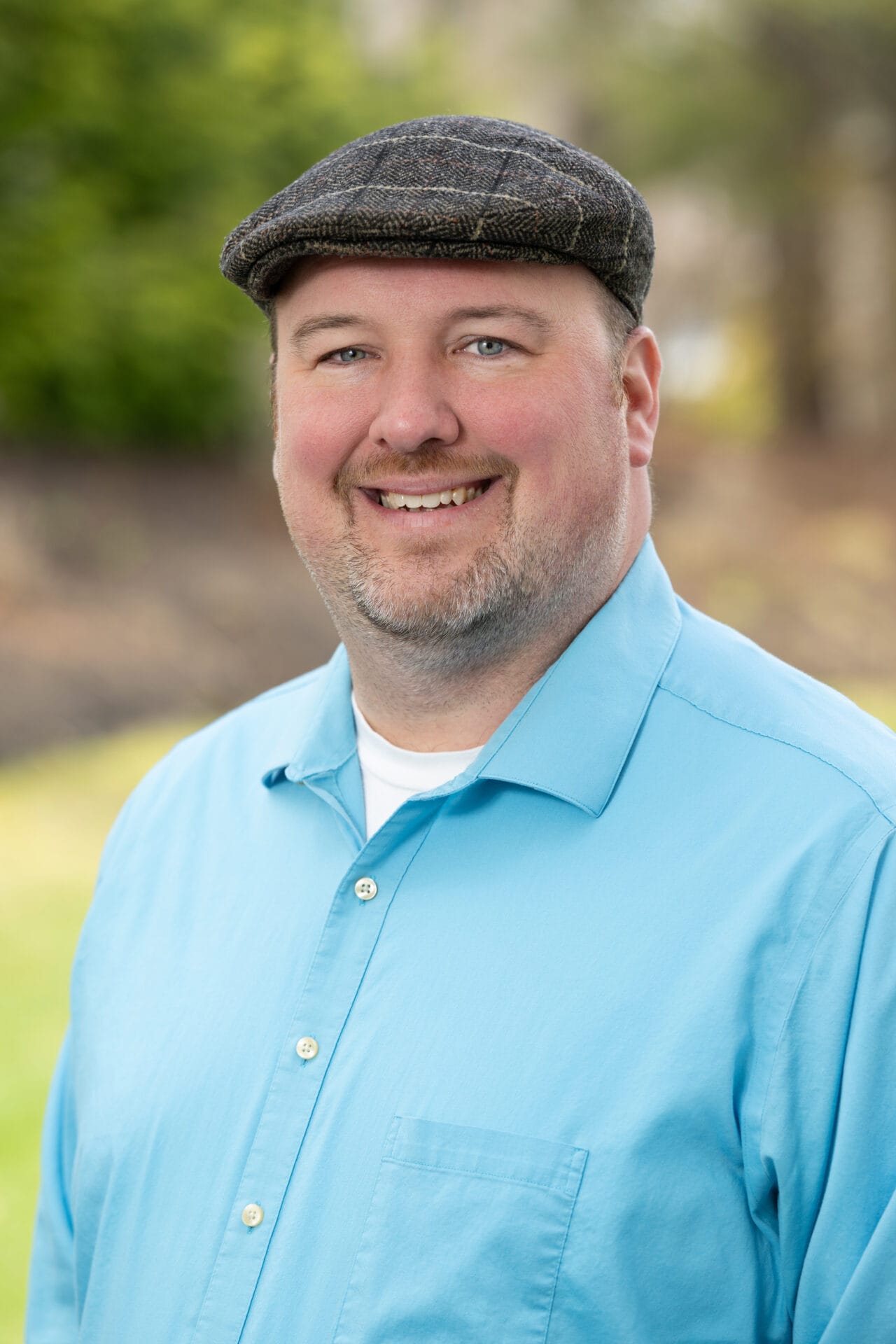 Man wearing a flat cap and a light blue button-up shirt, smiling outdoors with a blurred background of greenery.