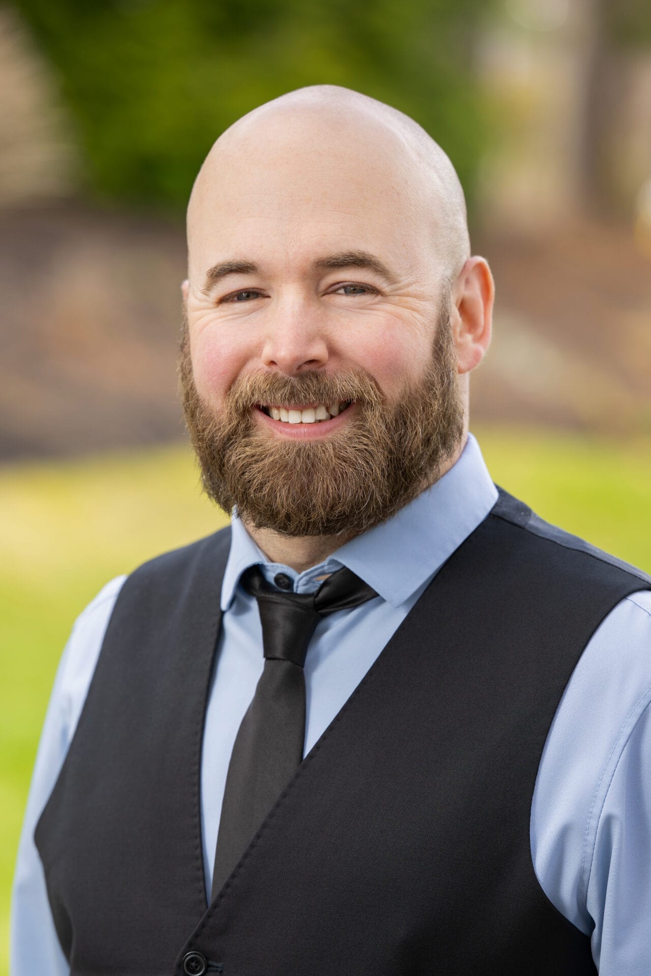 A bearded, bald man in a light blue shirt, black vest, and tie smiles outdoors with blurred greenery in the background.