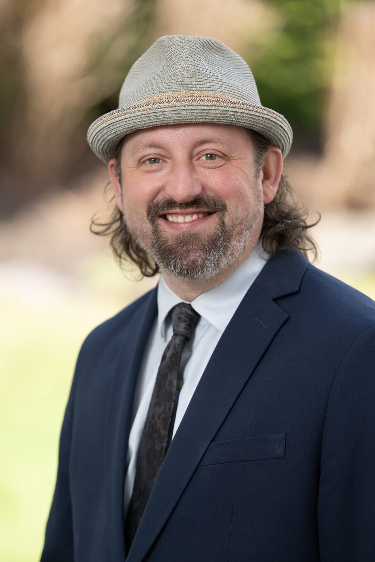 Man with long hair and beard wearing a light-colored fedora, navy suit, white shirt, and patterned tie, smiling outdoors with a blurred background.