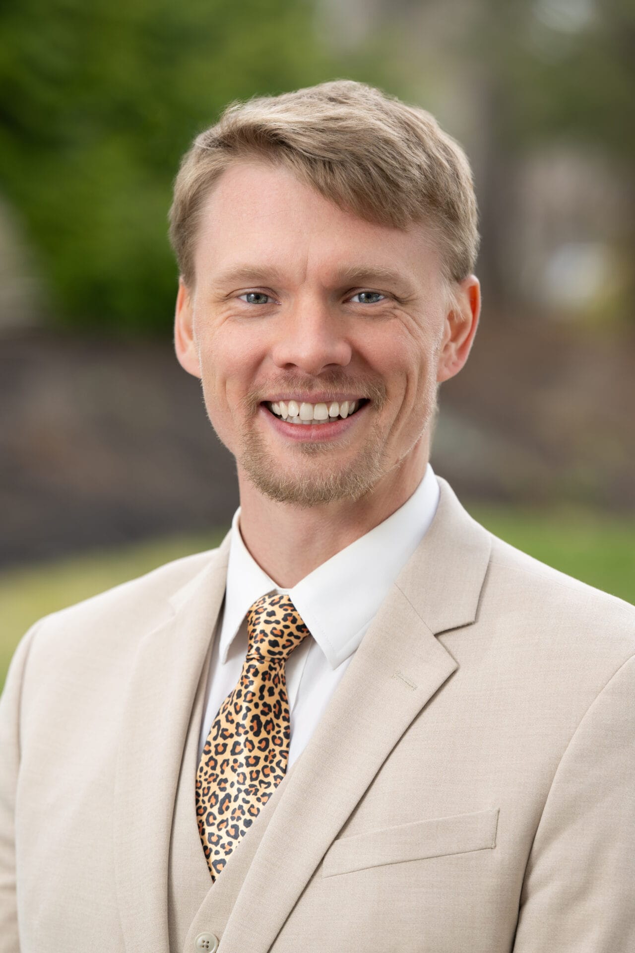 A man in a beige suit and leopard print tie smiles outdoors, with a blurred natural background.