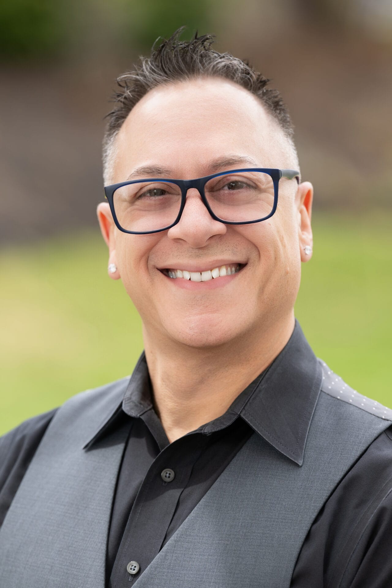 A person with short dark hair, glasses, and earrings smiles at the camera while wearing a black shirt and gray vest, outdoors with a blurred green background.