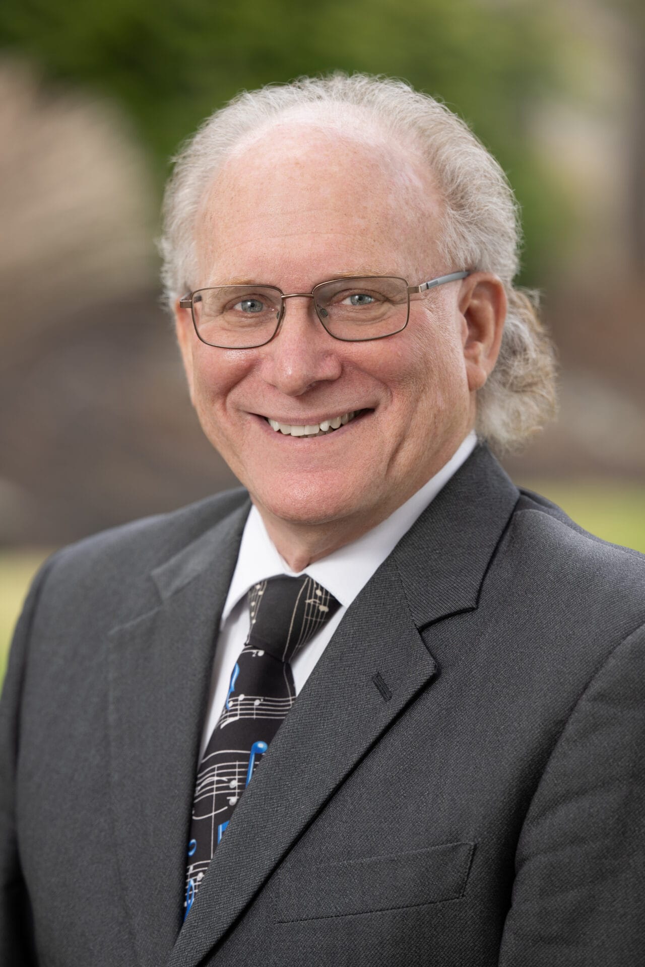Smiling older man with glasses and gray hair wearing a dark suit, white shirt, and black tie with a music notes pattern, standing outdoors.