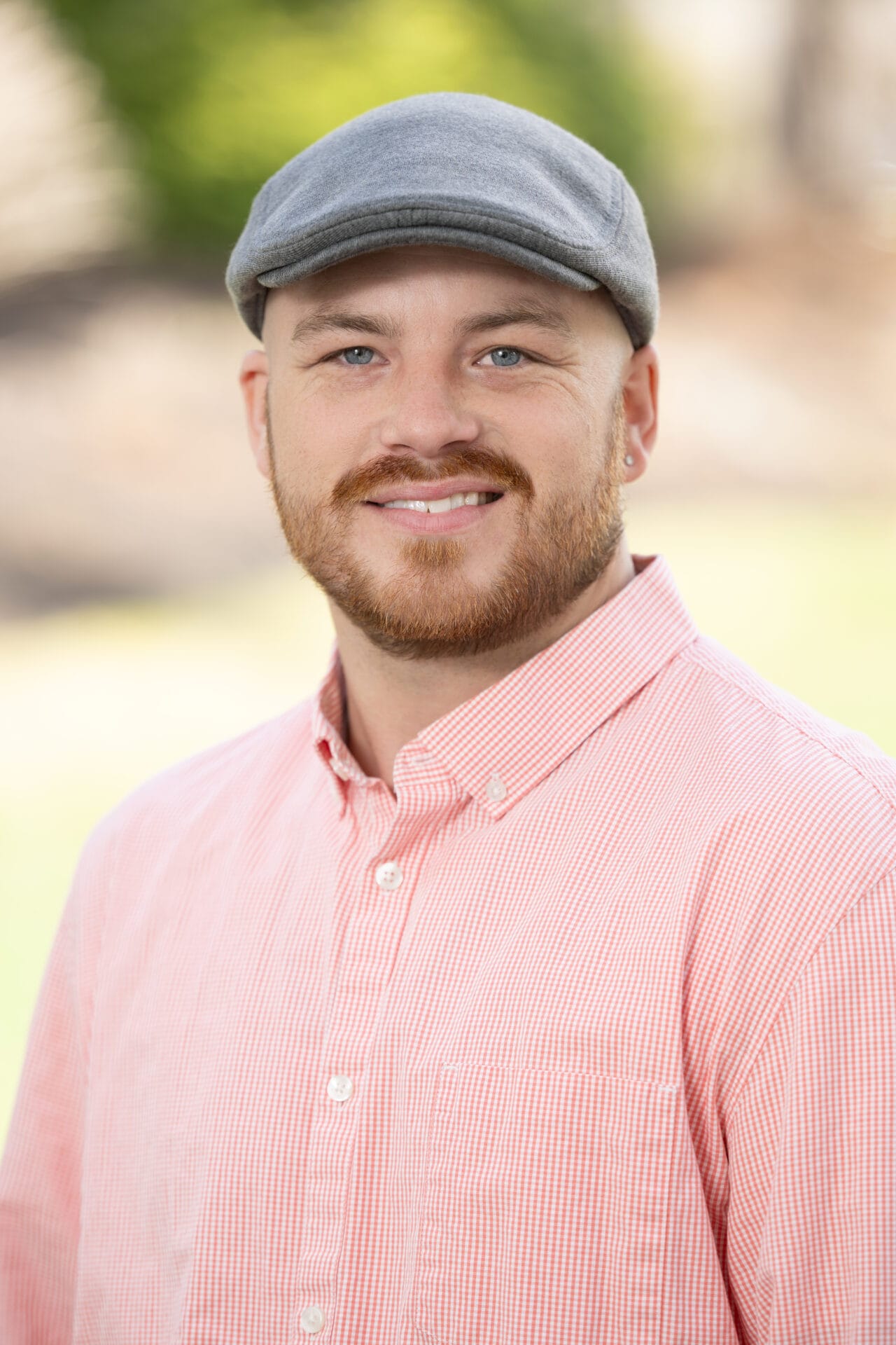 Man with a trimmed beard wearing a gray flat cap and light red checkered shirt, standing outdoors with a blurred background.