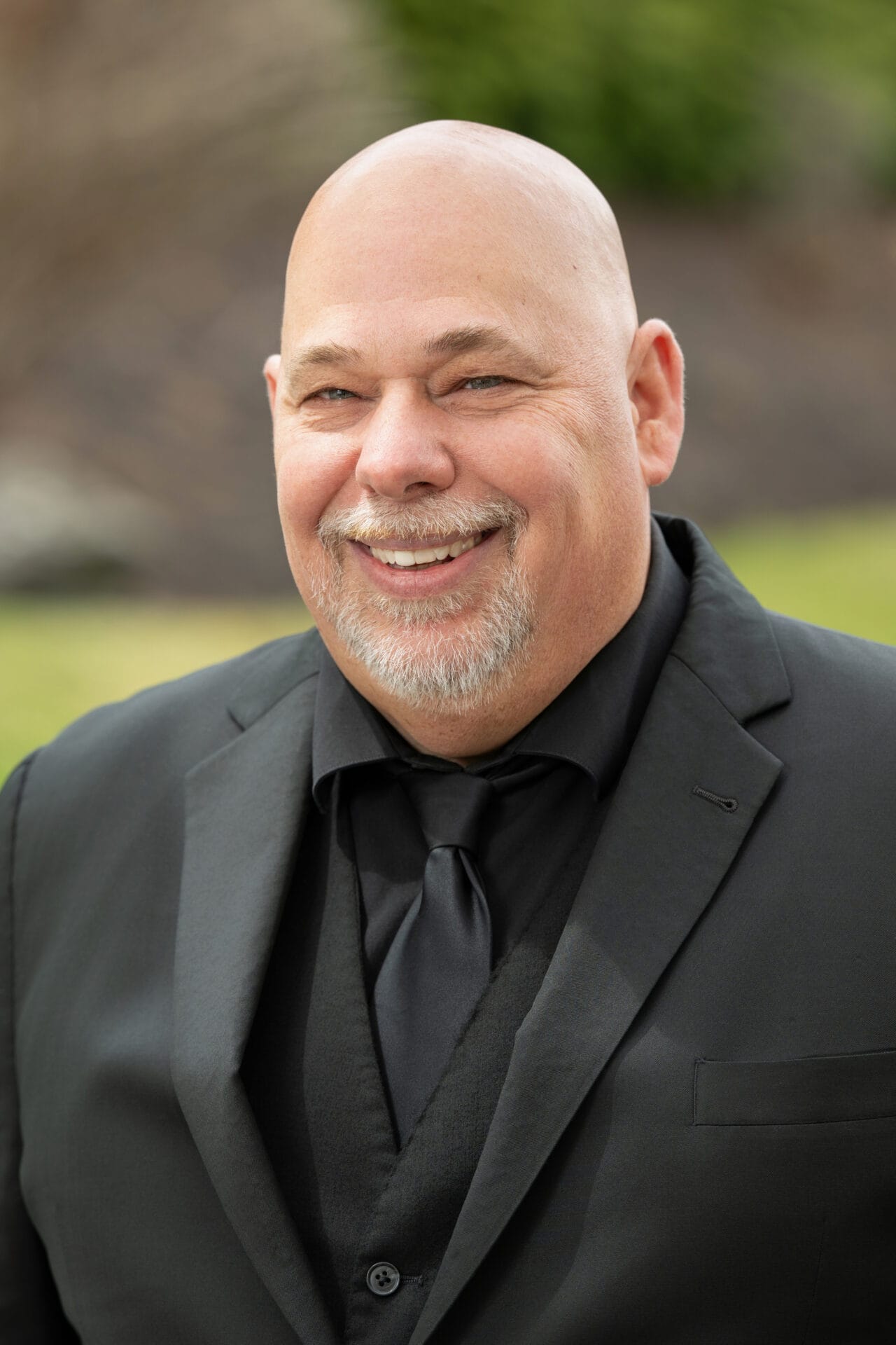 Bald man with a gray goatee, wearing a black suit and tie, smiles outdoors with blurred greenery in the background.