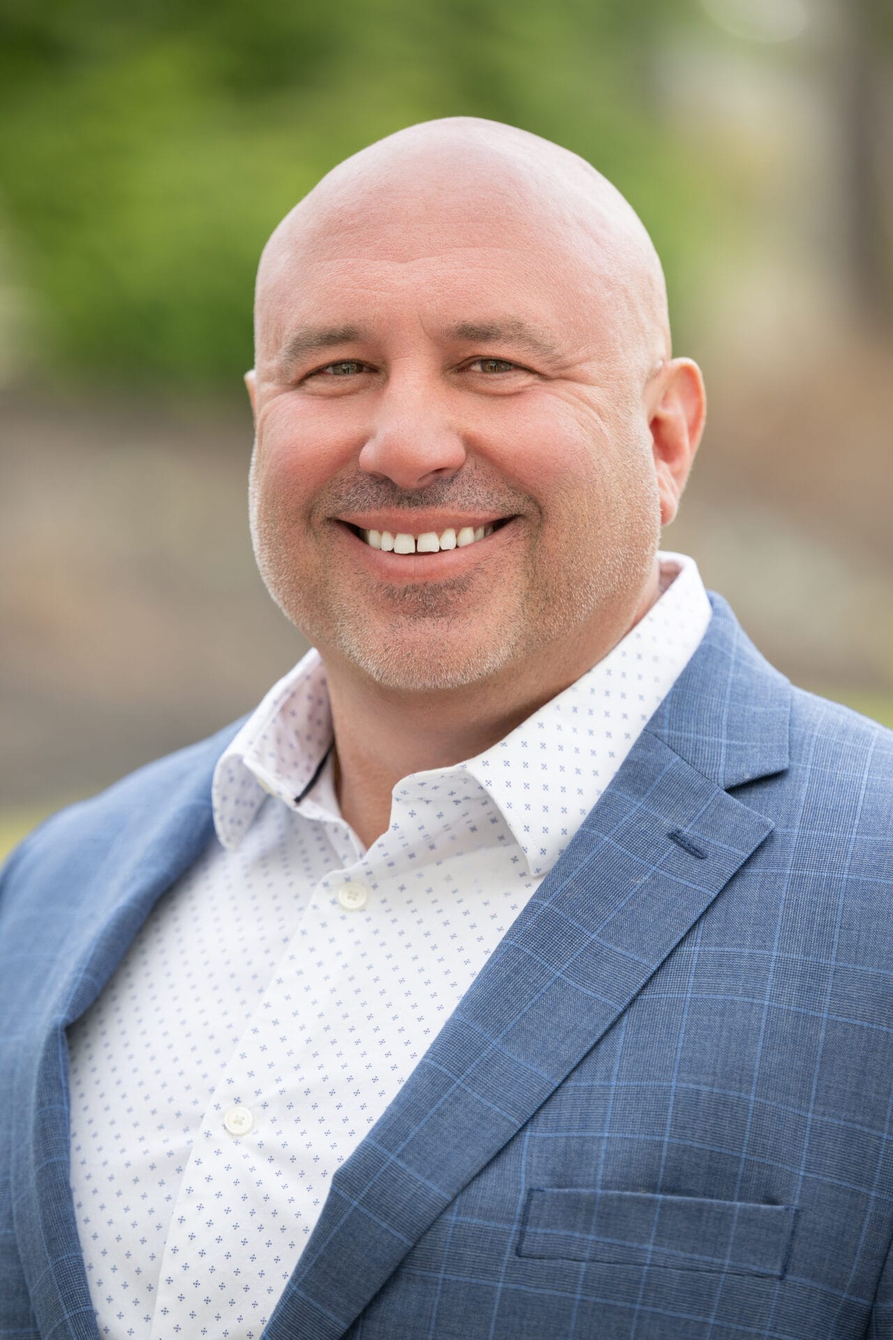 Bald man in a blue plaid suit jacket and white dress shirt smiling outdoors, with a blurred green background.