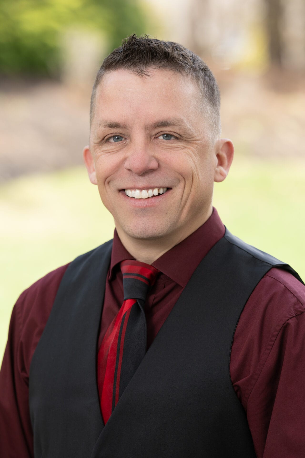 Smiling man with short brown hair wearing a maroon shirt, black vest, and red-and-black striped tie, standing outdoors with blurred greenery in the background.