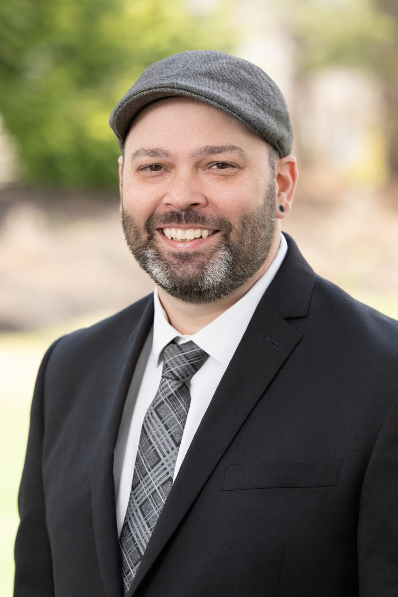 Man with a beard wearing a gray flat cap, black suit, white shirt, and patterned tie, smiling outdoors with a blurred green background.