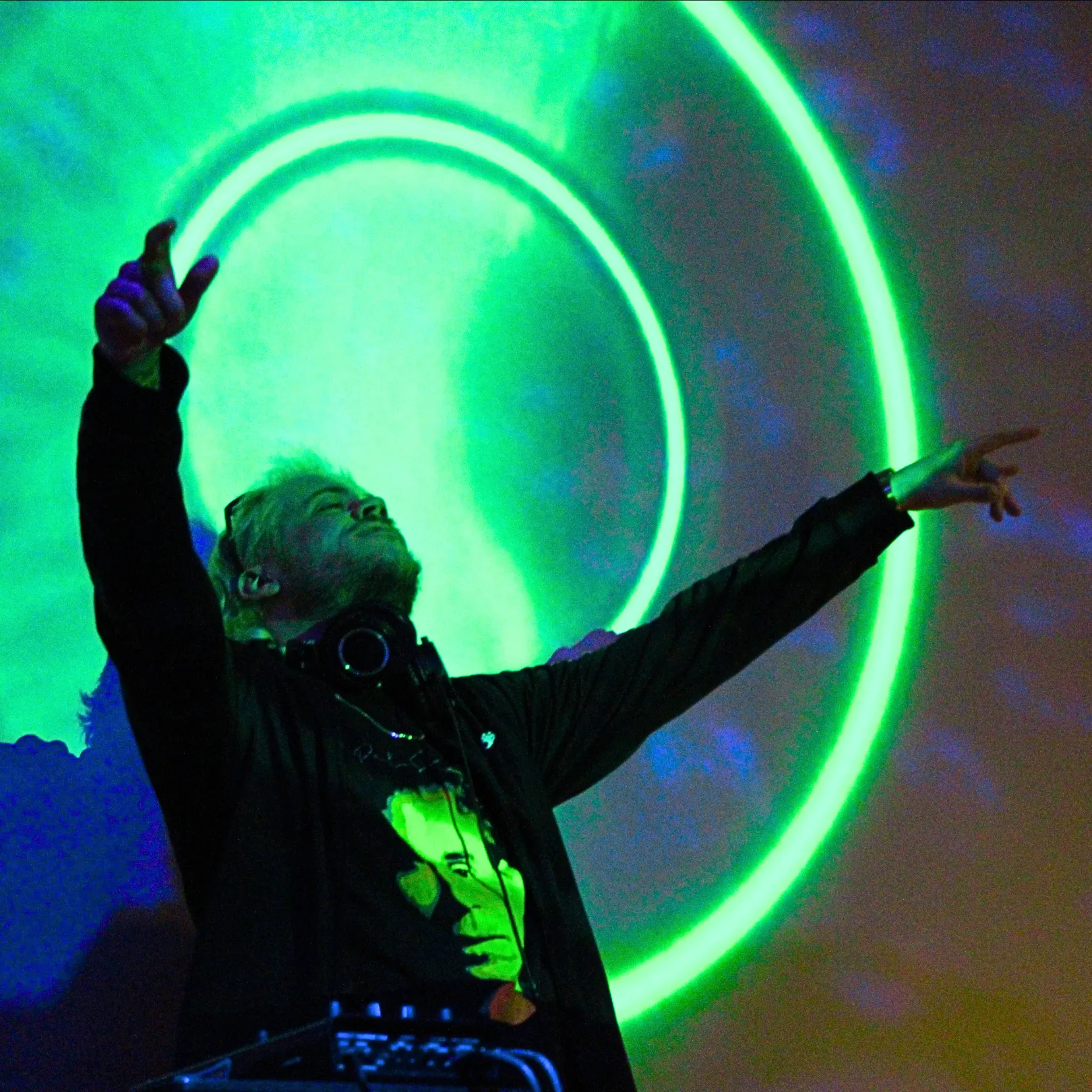 A DJ stands with arms raised in front of a green circular light pattern, wearing headphones and a graphic t-shirt, performing at an event.
